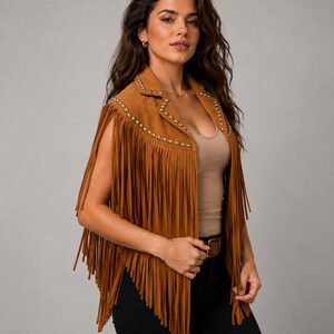 May include: A tan suede vest featuring long fringe detailing along the hem and arm openings, accented with a row of small metallic studs lining the lapels and front edges, worn over a neutral-colored tank top and black pants.