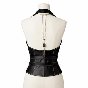 May include: Black leather corset with a halter neckline and a structured silhouette. The corset features a series of horizontal straps and decorative stitching. A silver-toned necklace with a cross pendant and a square charm hangs down the back.