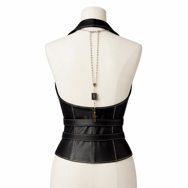 May include: Black leather corset with a halter neckline and a structured silhouette. The corset features a series of horizontal straps and decorative stitching. A silver-toned necklace with a cross pendant and a square charm hangs down the back.