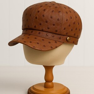 May include: A brown leather newsboy cap with a rounded crown and a short, curved brim. The cap features a pattern of small, evenly spaced dark brown dots. It is displayed on a mannequin head with a wooden stand.