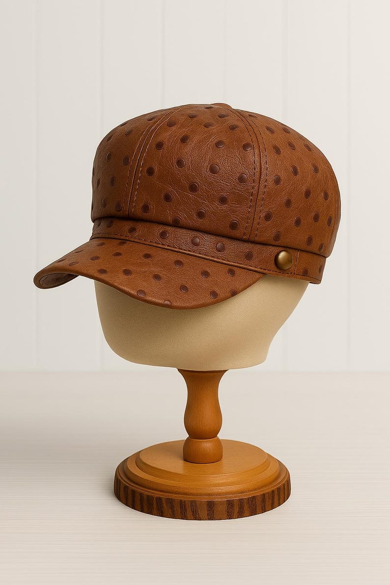 May include: A brown leather newsboy cap with a rounded crown and a short, curved brim. The cap features a pattern of small, evenly spaced dark brown dots. It is displayed on a mannequin head with a wooden stand.
