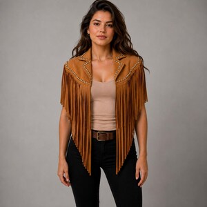 May include: A tan suede vest featuring long fringe detailing along the front panels and shoulders. The garment includes a notched collar accented with small silver-toned metal studs along the lapels and a sleeveless design.