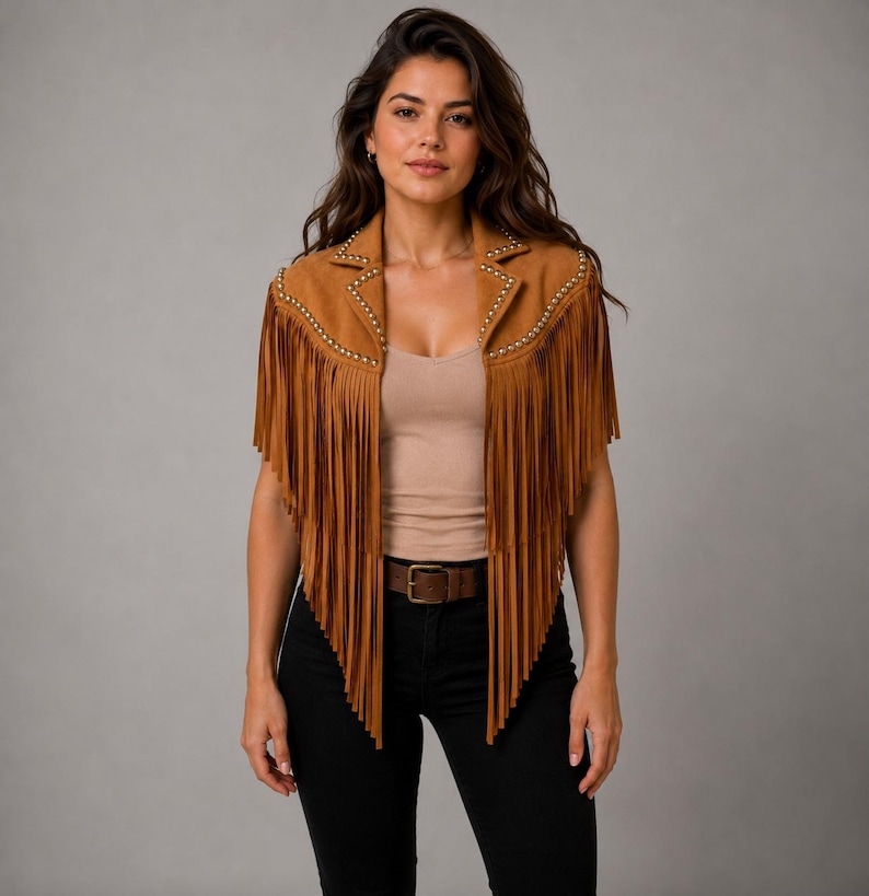 May include: A tan suede vest featuring long fringe detailing along the front panels and shoulders. The garment includes a notched collar accented with small silver-toned metal studs along the lapels and a sleeveless design.