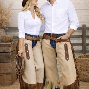 May include: A couple wearing cowboy hats, white button-down shirts, blue jeans, and tan leather chaps with fringe. One person holds a rope. The background includes wooden crates and a fence.