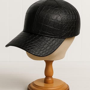 May include: Black baseball cap with a textured, crocodile-like pattern. The cap is displayed on a wooden hat stand with a round base. The hat has a curved brim and a button on top.
