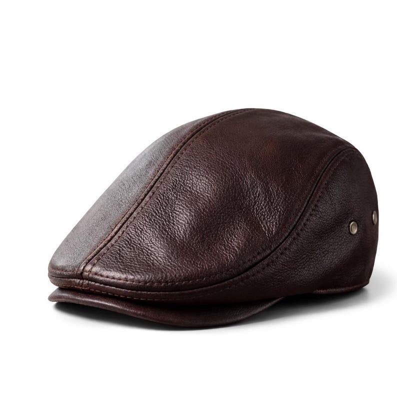 May include: A brown leather flat cap with a rounded crown and a small, stiff brim. The cap has a textured surface and is secured with two small metal rivets on the side. The cap is a classic style.