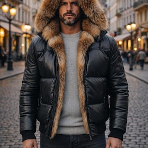May include: Black leather puffer jacket with a fur-lined hood. The jacket has a brown fur trim and a black ribbed hem and cuffs. The person is wearing a gray sweater and dark wash jeans. The background is a cobblestone street.