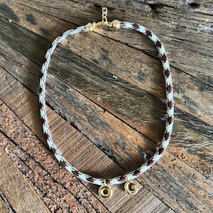 Puede incluir: Un collar con un cordón estampado en blanco y marrón. El collar tiene cierres dorados y dos colgantes dorados. El collar se muestra sobre una superficie de madera rústica.