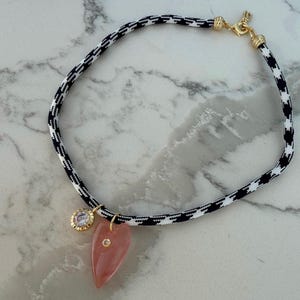 Puede incluir: Un collar con un cordón con estampado de pata de gallo en blanco y negro. El collar presenta un cierre dorado y dos colgantes: un colgante rosa en forma de corazón y un colgante redondo con una piedra transparente.