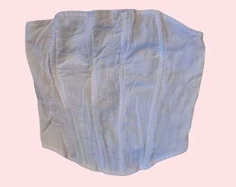 Puede incluir: Top de corsé blanco con bordado floral. La parte superior presenta un diseño estructurado con paneles verticales y un dobladillo curvo. La tela tiene un brillo sutil y el bordado agrega una textura delicada. El corsé es una prenda ligera y aireada.