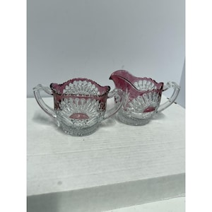 Puede incluir: Un juego de azucarero y crema de cristal transparente con un diseño decorativo de abanico. Los bordes son de color rosa intenso. El azucarero tiene dos asas y la crema tiene un pico. El juego es probablemente vintage.
