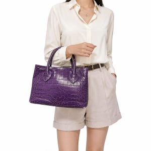 May include: A deep purple handbag featuring a textured crocodile-embossed pattern. The bag has two sturdy top handles, a rectangular silhouette, and a structured design with visible stitching along the base of the handles and the main body.