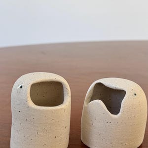 May include: Two speckled ceramic containers with rounded shapes and openings. One has a rectangular opening, the other a curved opening. Each has a small black dot, possibly an eye. The objects are on a wooden surface.