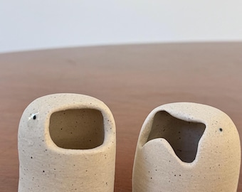 May include: Two speckled ceramic containers with rounded shapes and openings. One has a rectangular opening, the other a curved opening. Each has a small black dot, possibly an eye. The objects are on a wooden surface.