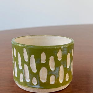 May include: A small, handmade ceramic cup with a green exterior and a cream-colored interior. The green surface features a pattern of irregular, white, oval-shaped cutouts. The cup sits on a wooden surface.