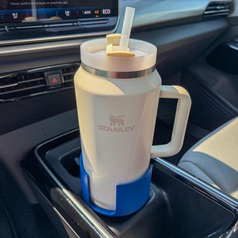 Puede incluir: Un termo de viaje Stanley blanco con asa y pajita, en un portavasos azul de coche. El termo tiene un borde plateado y una tapa beige. El logotipo de Stanley es visible.