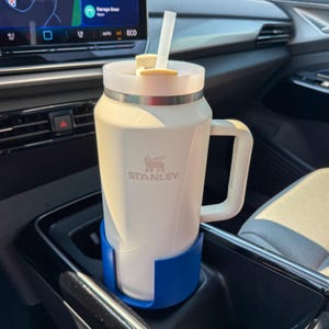 Puede incluir: Un termo de viaje blanco Stanley con asa y pajita, en un portavasos azul en un coche. El termo tiene una banda plateada y el logotipo de Stanley. Capacidad aproximada de 0,7 litros.