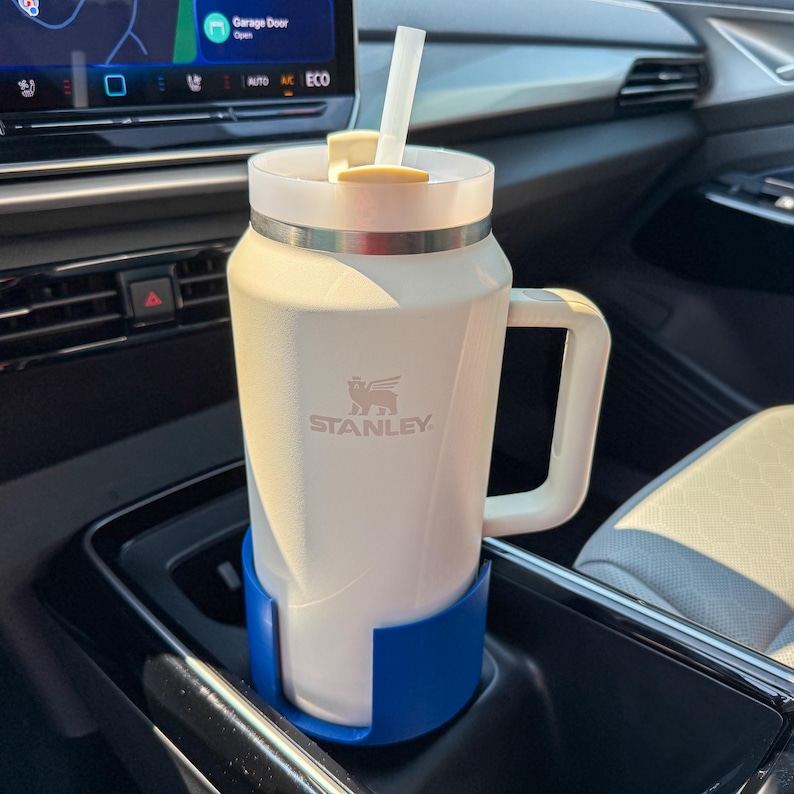 Puede incluir: Un termo de viaje blanco Stanley con asa y pajita, en un portavasos azul en un coche. El termo tiene una banda plateada y el logotipo de Stanley. Capacidad aproximada de 0,7 litros.