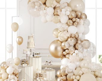 May include: An elegant display featuring a tiered cake, cupcakes, and other desserts on pedestals. A large balloon arch in shades of gold, white, and clear balloons frames the scene. Additional balloons in similar colors are scattered around the display.
