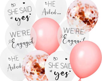 May include: A collection of balloons in white, solid pink, and clear with rose gold confetti. The white balloons feature black text reading "SHE SAID yes", "HE Asked...", and "WE'RE Engaged" along with small ring and heart icons printed on the surface.