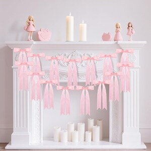 May include: A decorative garland featuring a series of light pink satin ribbon bows arranged in a cascading pattern. The bows are tied with long, flowing tails and are connected by a thin string, creating a uniform display of soft pink fabric.