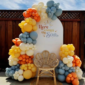 May include: An arched white backdrop featuring the text Here comes the Son with a sun graphic, decorated with a large balloon garland in shades of orange, yellow, cream, and blue, positioned behind a light brown woven rattan peacock chair.