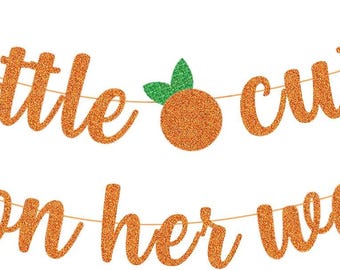 May include: A glittery orange banner with the text "a little cutie is on her way" in cursive script. The banner includes orange fruit designs with green leaves. The banner is strung on a thin, light brown string.