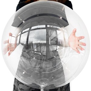 May include: A large, perfectly spherical, transparent balloon made of clear plastic material. The surface of the balloon is highly reflective, creating a distorted, wide-angle view of a cityscape with buildings and windows visible through the sphere.