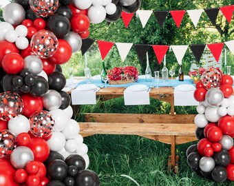 May include: A festive outdoor party scene with a wooden picnic table set for a celebration. A balloon arch in red, black, silver, and white decorates the scene. A banner with red, black, and white pennants hangs above the table, which is set with plates and flowers.