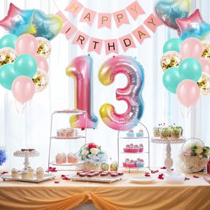 May include: A birthday party decoration set featuring a pink "HAPPY BIRTHDAY" banner, large iridescent number 13 balloons, and clusters of pastel pink, teal, and gold confetti balloons, arranged above a table with tiered dessert stands.