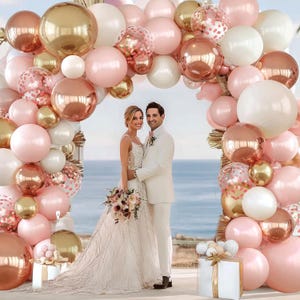 May include: A wedding scene featuring a couple standing under an arch of balloons. The balloons are in shades of gold, rose gold, pink, and white. The bride wears a white gown and holds a bouquet. The groom is in a white suit. The background is the ocean.