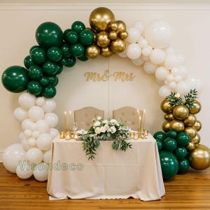 May include: A wedding reception table is decorated with a beige tablecloth, candles, and floral arrangements. A balloon arch in green, gold, and white surrounds the table. The words "Mr & Mrs" are displayed above the table.