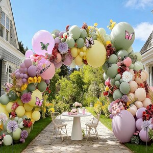 May include: A large balloon arch featuring a mix of pastel pink, sage green, yellow, and lavender balloons. The arrangement is decorated with artificial flowers and butterfly cutouts, creating a decorative frame over a white table and chairs.