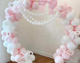 Pode incluir: Um arco decorativo feito de balões brancos e rosa, adornado com fios de pérolas brancas e laços rosa. O arco é suportado por uma estrutura de metal dourado, perfeito para um fundo de festa. A palavra "Patecha" está escrita no arco.