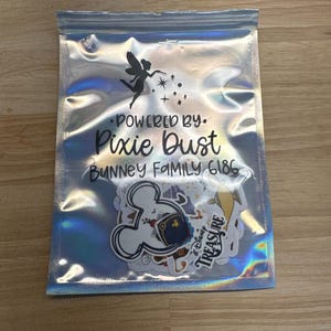 May include: A clear, iridescent bag containing stickers. The bag has text that reads "Powered by Pixie Dust" and "Bunney Family 6186". The stickers feature cartoon characters and the word "Treasure".