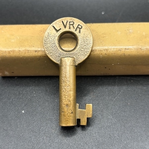 Könnte beinhalten: Ein antiker Messingschlüssel mit den Buchstaben "LVRR" auf einem kreisförmigen Kopf eingraviert. Der Schlüssel hat einen zylindrischen Schaft und einen rechteckigen Bart. Der Schlüssel ist vor einem dunklen Hintergrund platziert.