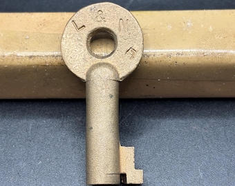 May include: A brass-colored metal key with a circular bow featuring the embossed letters L & N. The key has a cylindrical shank and a simple bit at the bottom. The surface of the metal has a matte, textured finish with visible vertical lines.