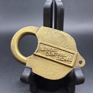 Puede incluir: Un candado de latón antiguo con un asa curva y un cuerpo rectangular. El texto "ILLINOIS CENTRAL SIGNAL" está grabado en relieve en la parte delantera. El candado se exhibe sobre un soporte negro sobre un fondo oscuro.