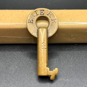 Puede incluir: Una llave de latón antigua con el texto "ERIE RR" en un diseño circular en la parte superior. La llave tiene un vástago rectangular y una paleta distintiva. La llave está sobre un fondo neutro.