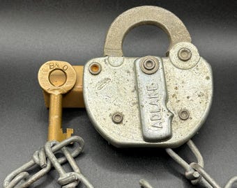May include: A weathered silver-toned Adlake padlock with a rounded shackle, featuring a vertical metal plate embossed with the brand name. A brass key stamped with B&O is positioned beside the lock, which is attached to a heavy metal chain.
