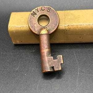 May include: A vintage brass-toned metal key with a circular bow featuring the stamped letters N.Y.C.S. The key has a cylindrical shank and a simple bit pattern at the end, showing signs of oxidation and wear on its metallic surface.