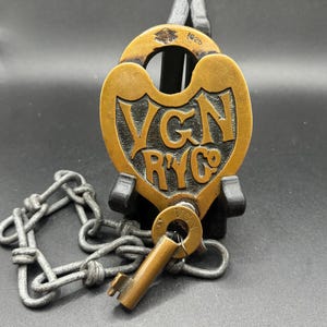 Puede incluir: Un candado antiguo de latón con llave y cadena. El candado tiene grabadas las letras "VGN RYC Co" y el año "1925". La llave está insertada en la cerradura. La cadena es de metal.