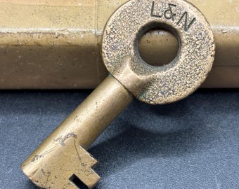 May include: A weathered brass key featuring a round bow with the stamped letters L&N. The key has a cylindrical shank and a notched bit at the end, showing signs of age and oxidation on its metallic surface, with a textured finish on the bow.