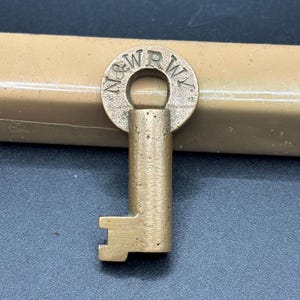 Puede incluir: Una llave de latón vintage con las letras "N&WRY" estampadas alrededor de la cabeza circular. La llave tiene un vástago rectangular y una paleta dentada. La llave es probablemente una antigüedad o un objeto de colección.
