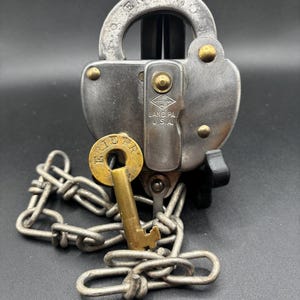 Puede incluir: Candado Erie Railroad vintage con llave y cadena. El candado es de metal plateado con remaches dorados y una llave. La llave está insertada en el candado. El texto "ERIE RR" es visible en el candado.