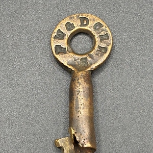 May include: Antique brass key with a circular top and a cylindrical stem. The top is engraved with the letters "W & D G R" around a central hole. The key shows signs of age and wear, with a slightly damaged bit.