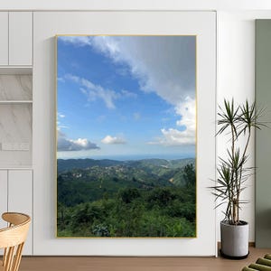 May include: A framed landscape art print depicting a mountain vista under a bright blue sky with white clouds. The artwork shows verdant green hills, a distant town, and a gold frame, creating a tranquil and natural aesthetic.