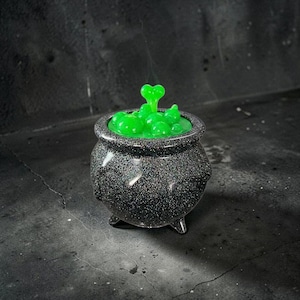 May include: A black cauldron with a glittery finish and three small feet, filled with a bright green liquid substance that features several raised, rounded bubbles and a central heart-shaped element protruding from the top surface.