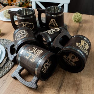 Könnte beinhalten: Eine Sammlung dunkler Bierkrüge aus Holz mit Edelstahlrändern und Griffen. Jeder Krug ist mit einem Namen und dem Wort "Groomsman" sowie einem Datum graviert. Die Krüge sind auf einer Holzoberfläche angeordnet.