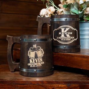 May include: Dark brown wooden beer mugs with metal bands and handles. One mug features a large letter "K" and the text "KEVIN GROOMSMAN 10.10.2025". Another mug has an image of two beer mugs clinking with the text "KEVIN GROOMSMAN 10.10.2025".